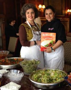 Two women in aprons holding cookbook by salads